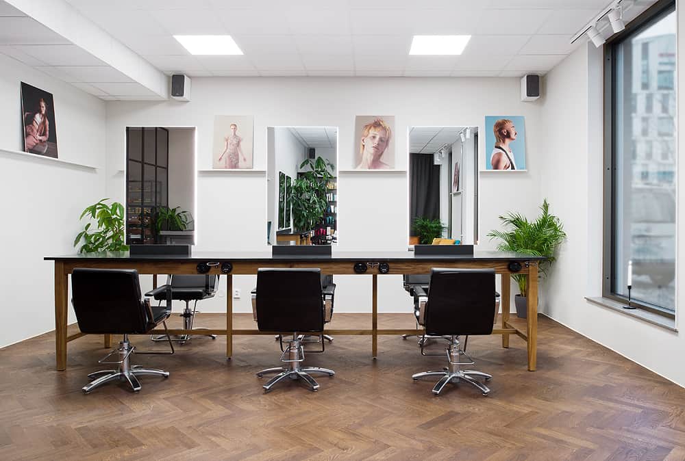 The picture shows a hair salon with four black barber chairs in front of a long wooden table with mirrors, decorated with portrait photographs on the wall above. The salon is bright and minimalist, with large windows, white walls and green plants giving the room a fresh feel.