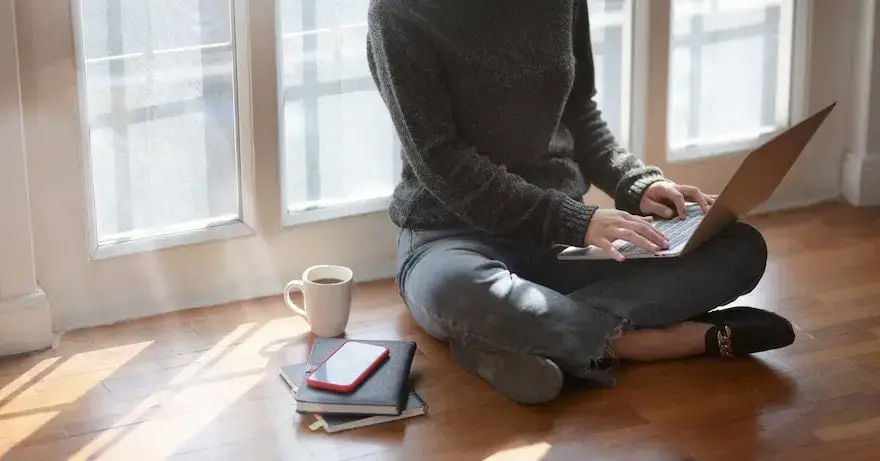 Person sitting on the floor by the window and typing on a laptop.