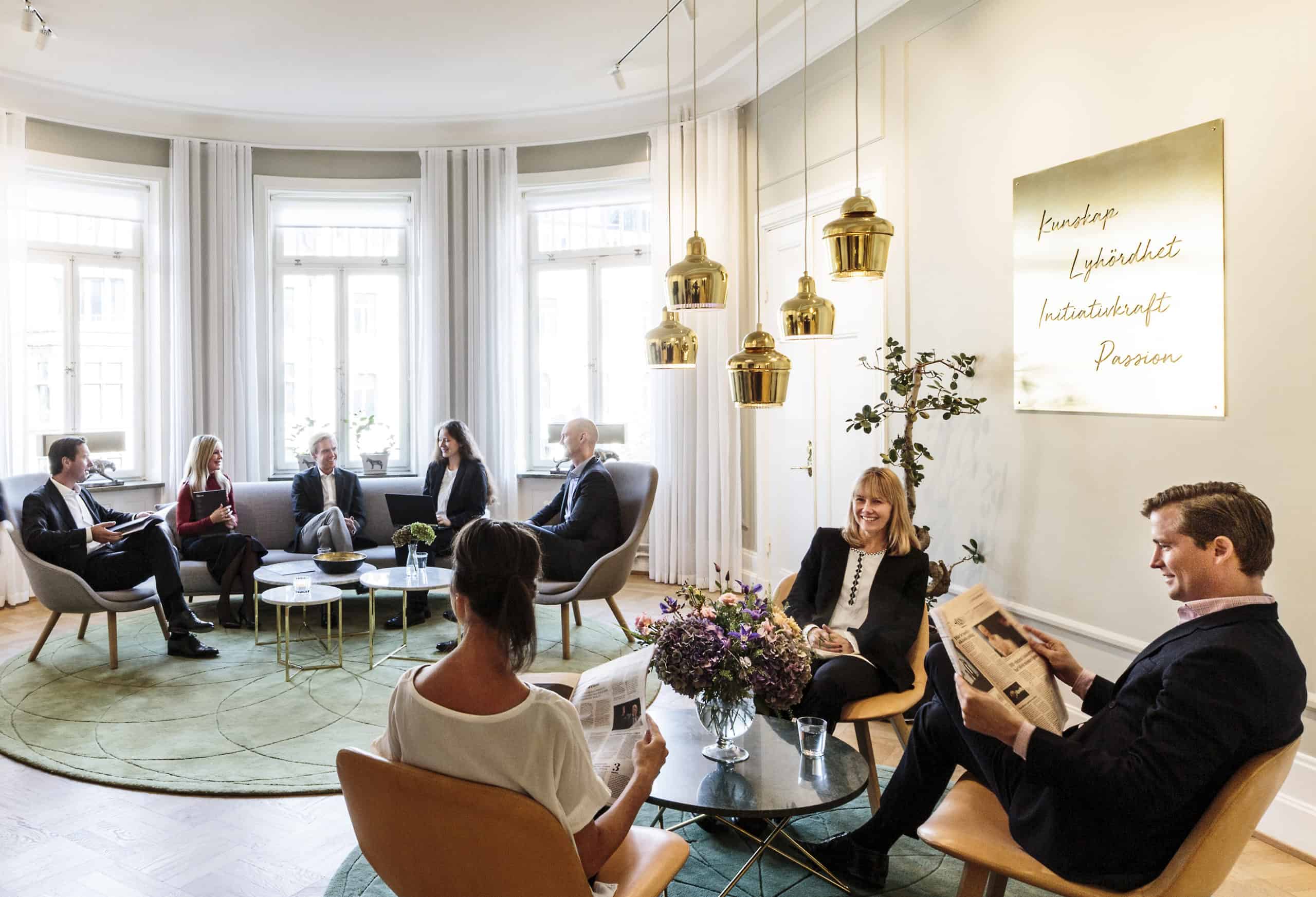 Pænt klædte mænd og kvinder sidder omkring to borde. En mand læser en avis.