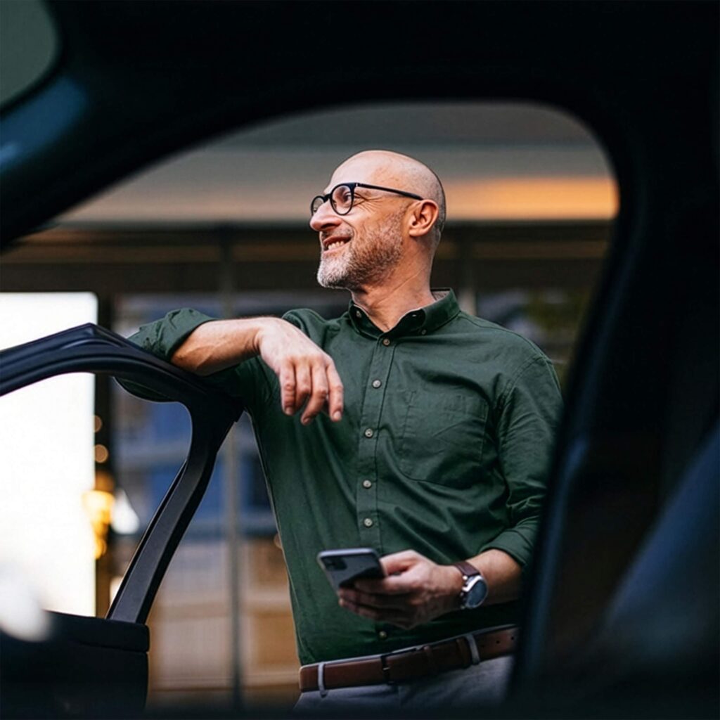 Man står med mobilen i handen vid en bil och blickar ut.